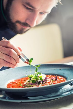 Chef in hotel or restaurant kitchen cooking, only hands. He is working on the micro herb decoration. Preparing tomato soup.